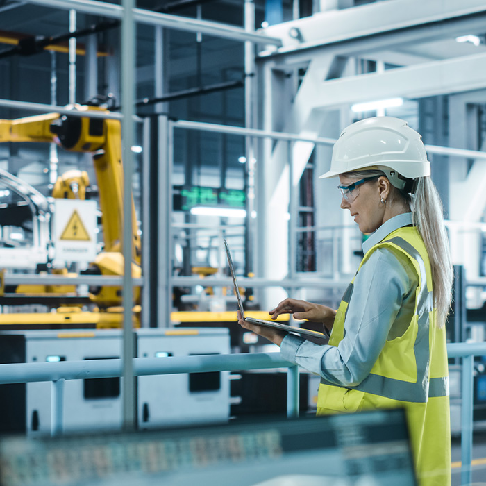 Female engineer in a high-vis vest and hard hat using a laptop in an industrial facility, representing support services for Flexible Energy Procurement.